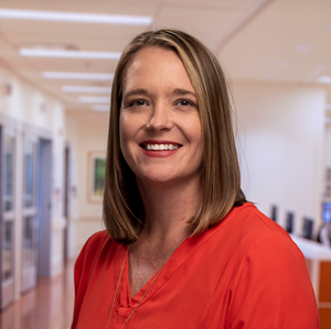 Portrait of Amy Hu, a blonde white woman in a red blouse. In the background is a blurry, but still recognizable, hospital hallway.