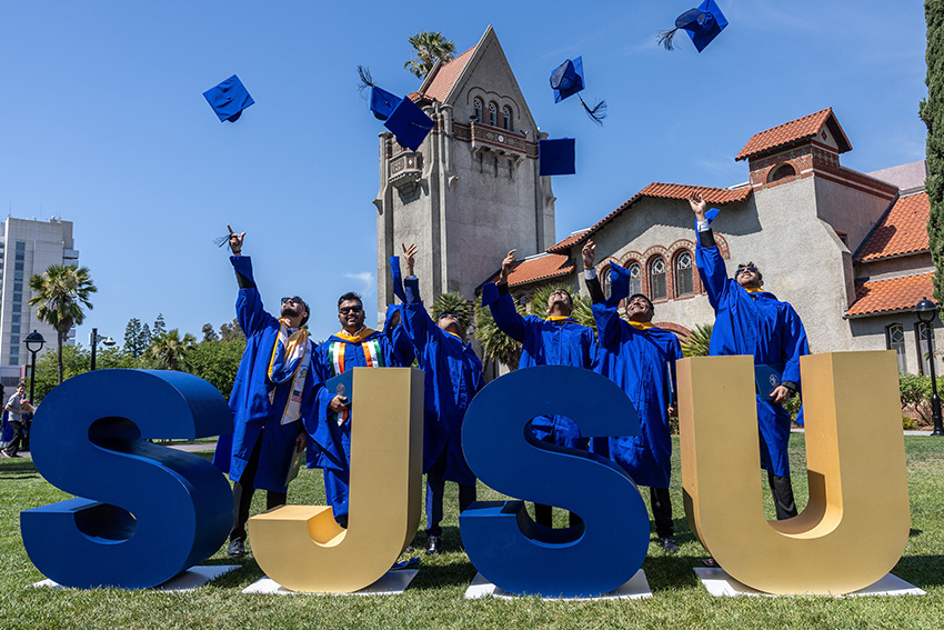 SJSU graduates toss their caps in the air while celebrating behind large SJSU letters on campus.